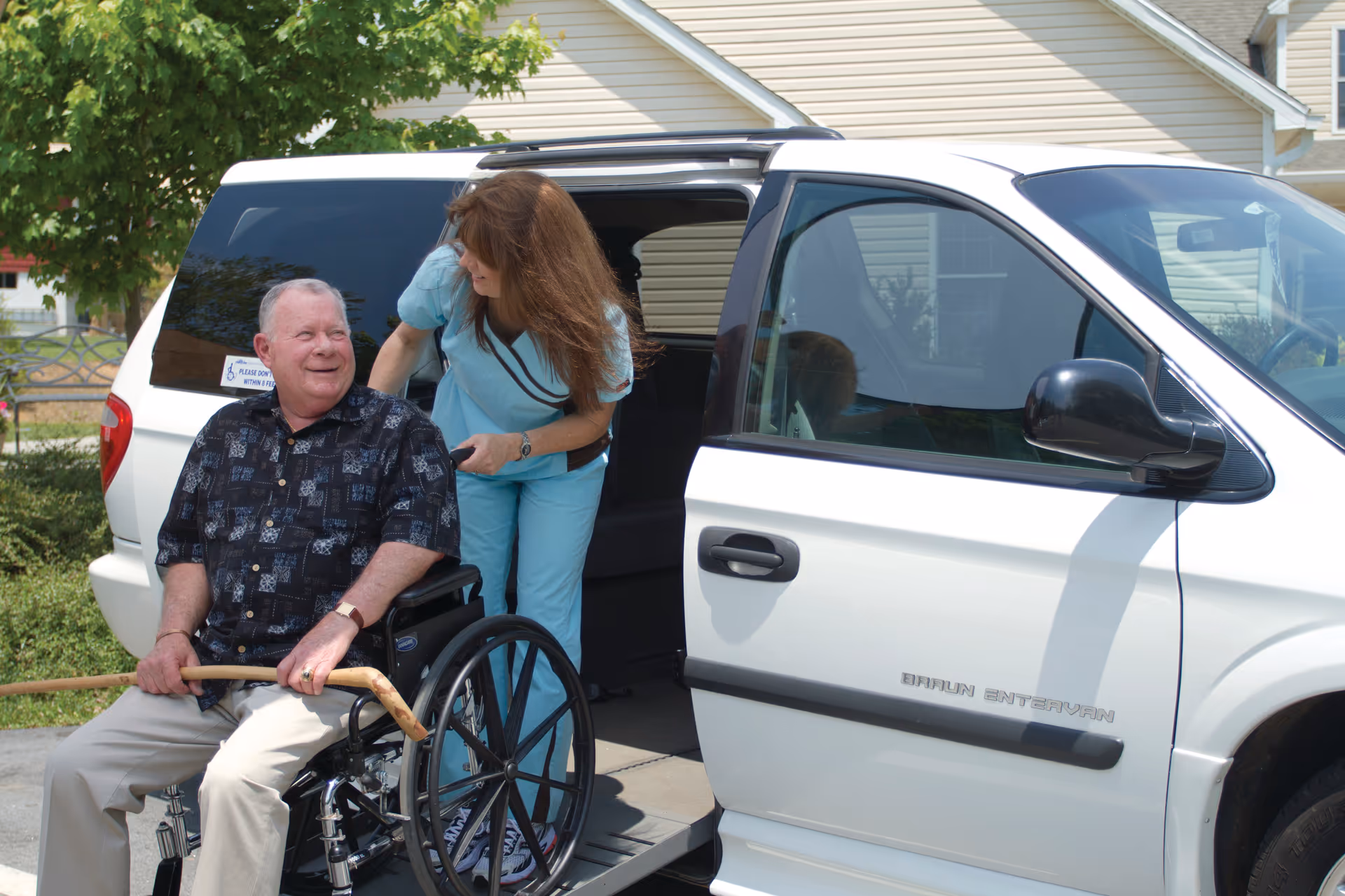 An elderly man in a wheelchair holding a wooden cane is being assisted by a female caregiver wearing light blue scrubs next to a white Braun Entervan with its side door open, outside a residential building with trees and bushes in the background.