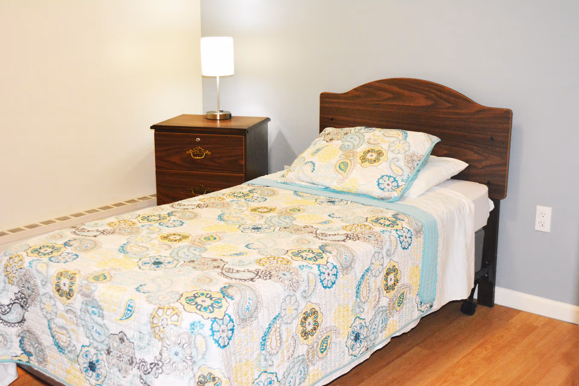 Single bed with a patterned quilt and pillow next to a wooden nightstand and lamp in a simple bedroom.