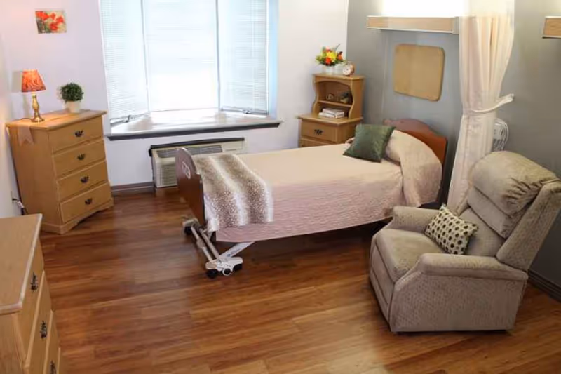 A senior living facility bedroom with a single adjustable bed covered with a beige blanket and a green pillow. There is a comfortable beige recliner chair with a patterned cushion beside the bed. The room has wooden flooring, a wooden dresser with a lamp and a small plant, a wooden nightstand with flowers and a clock, and a large window with blinds letting in natural light.