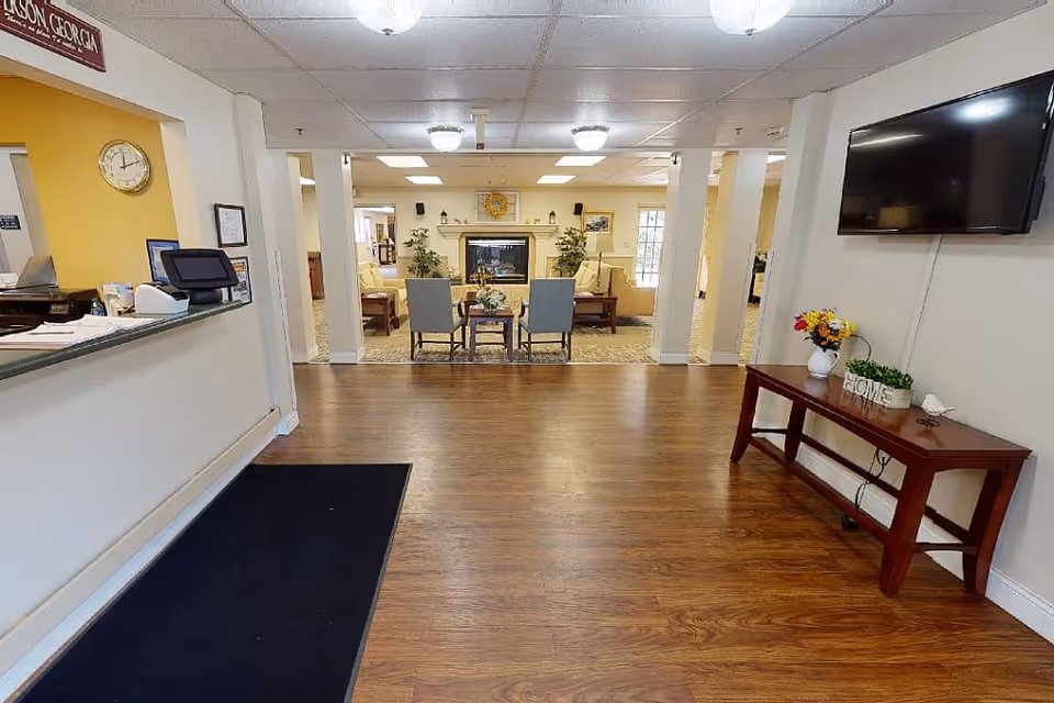 Interior view of a senior living facility lobby area with a reception desk on the left, a wooden floor, a black rug, and a seating area with chairs and a fireplace in the background. On the right wall, there is a mounted flat-screen TV above a wooden console table decorated with flowers and a small plant.