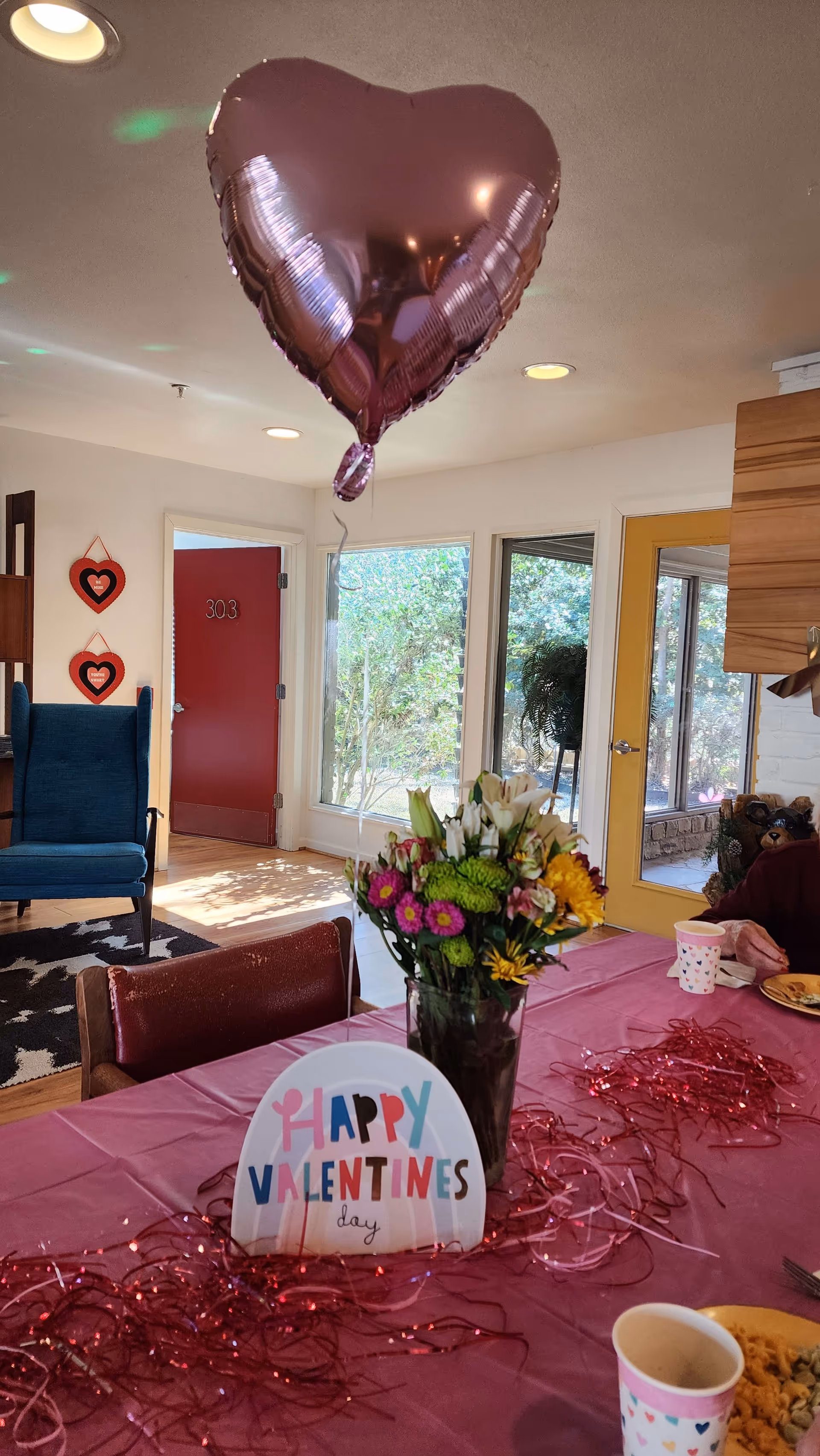 A decorated table with a pink tablecloth, a vase of colorful flowers, and a sign that says 'Happy Valentines day'. Above the table floats a large heart-shaped balloon. In the background, there is a blue armchair, a red door labeled 303, windows showing greenery outside, and a yellow door leading to a patio area.