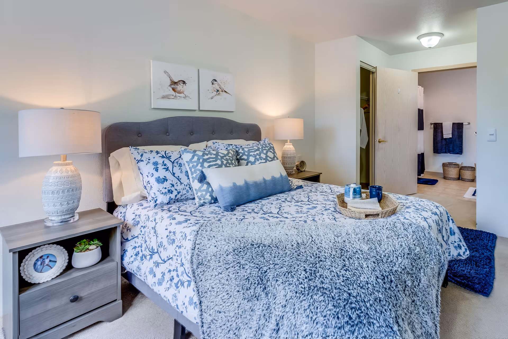 A cozy bedroom with a bed dressed in blue and white floral bedding, multiple decorative pillows, and a gray textured throw blanket. Two bedside tables each hold a lamp with a patterned base. Above the bed are two framed bird illustrations. An open door reveals a walk-in closet and a bathroom with blue towels and woven baskets.