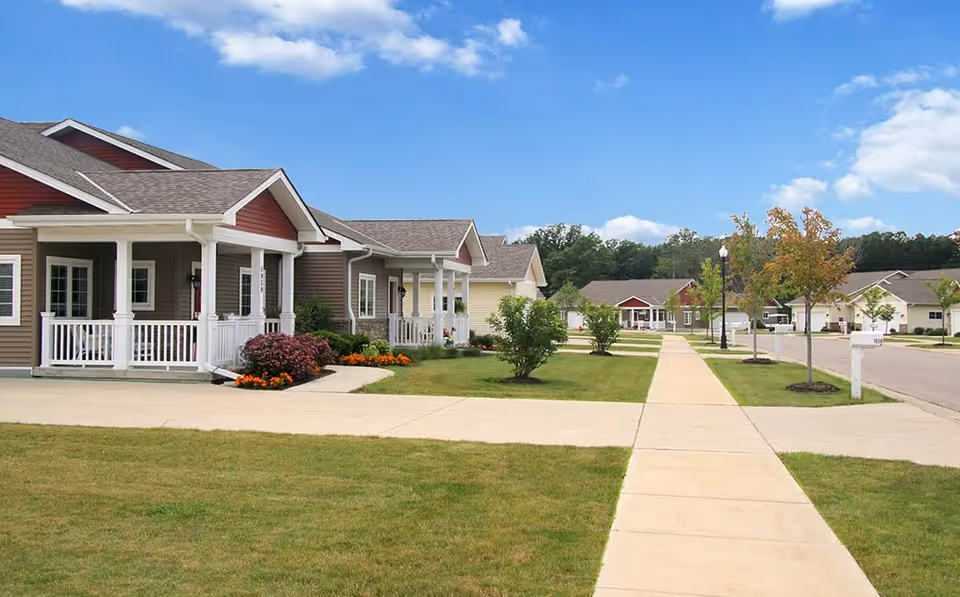 Row of single-story ranch-style homes with front porches lining a sidewalk and manicured lawns under a blue sky.