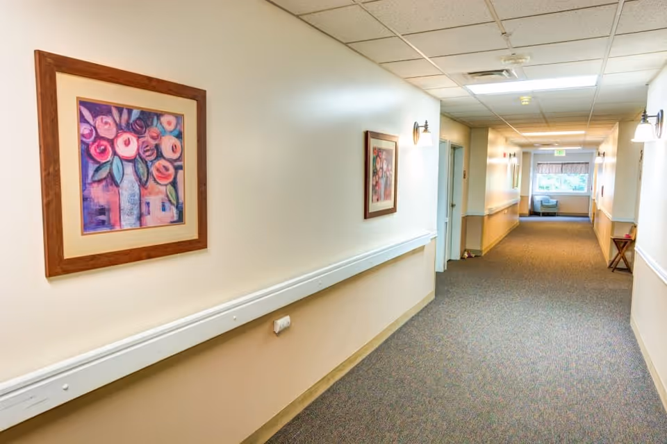 A well-lit hallway in a senior living facility with beige walls and carpeted floor. The walls are decorated with framed floral paintings and wall-mounted light fixtures. At the end of the hallway, there is a window with natural light and a light blue armchair beneath it.