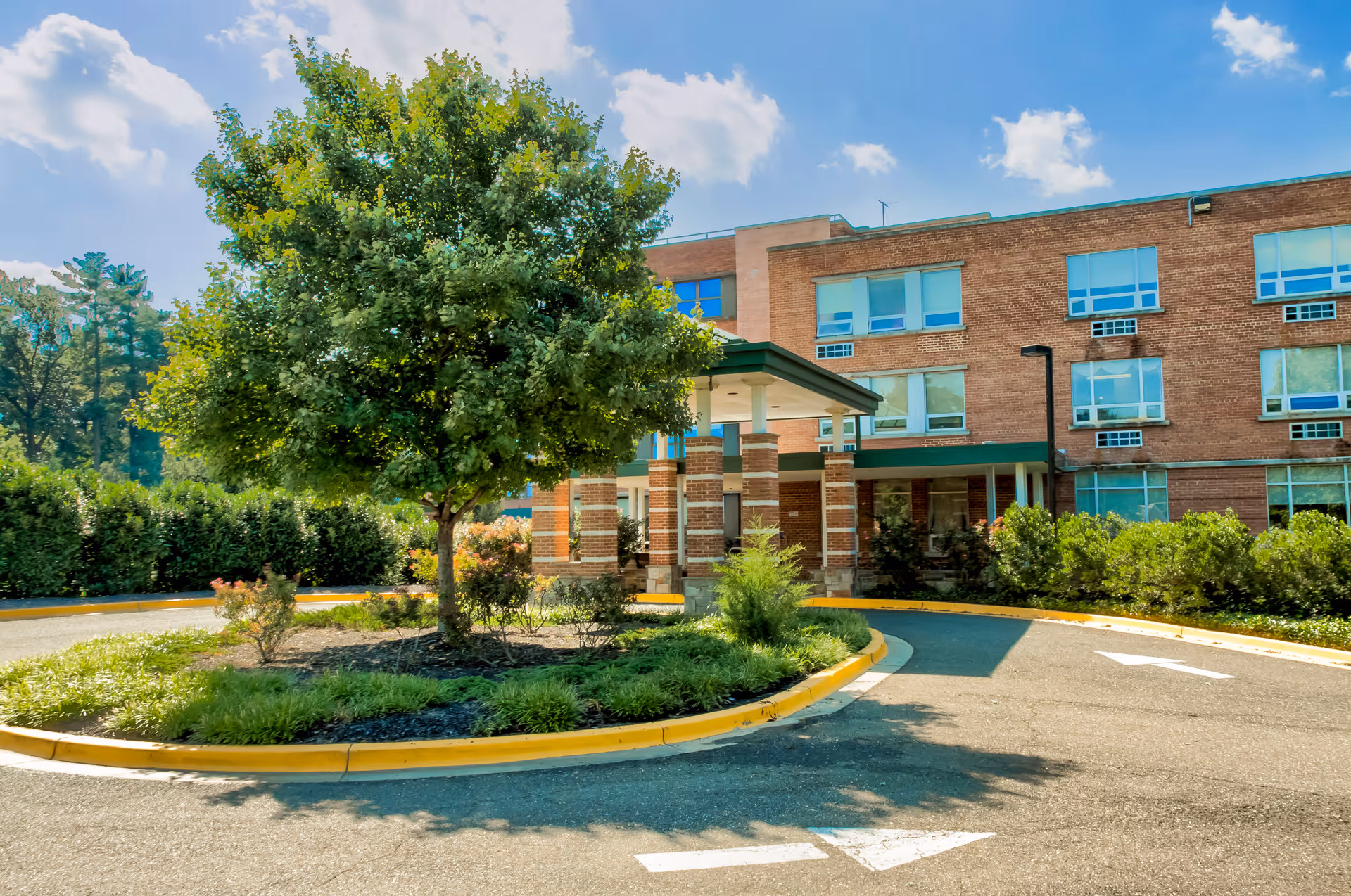 Exterior view of a brick senior living facility building with multiple windows and a covered entrance. A circular driveway with a landscaped island featuring a large tree and shrubs is visible in front of the entrance. The sky is partly cloudy with blue patches.