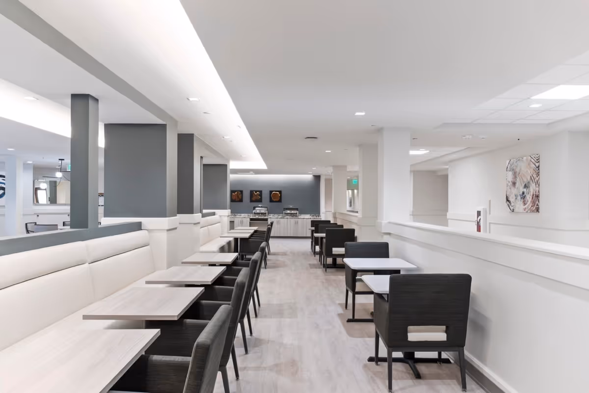 A modern dining room with light wood tables and dark cushioned chairs arranged in rows along a white cushioned bench and opposite wall. The room has light-colored flooring, white walls, and ceiling with recessed lighting. At the far end, there is a counter with serving trays and decorative wall art above it.