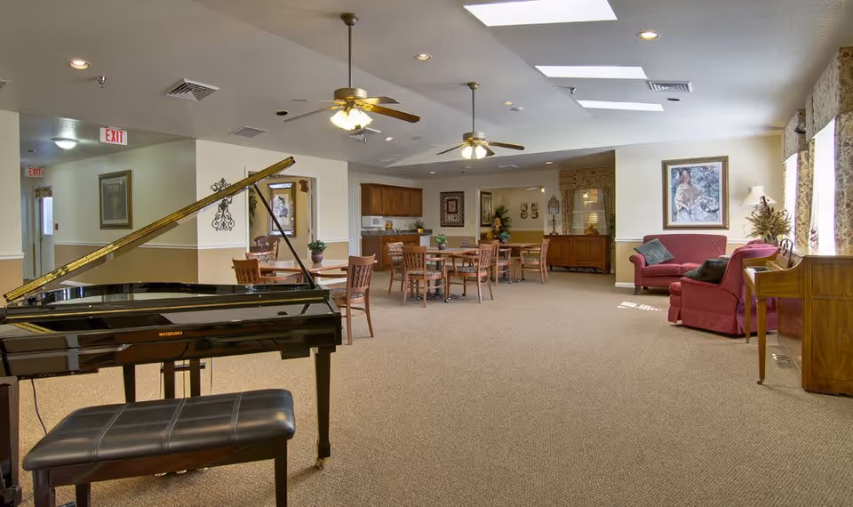 A spacious senior living common area featuring a black grand piano with a matching bench in the foreground. The room has beige carpeting, several wooden tables and chairs, two ceiling fans with lights, and a seating area with red upholstered armchairs and a sofa near large windows with floral curtains. The walls are decorated with framed artwork and plants, and there is a wooden cabinet along one wall.