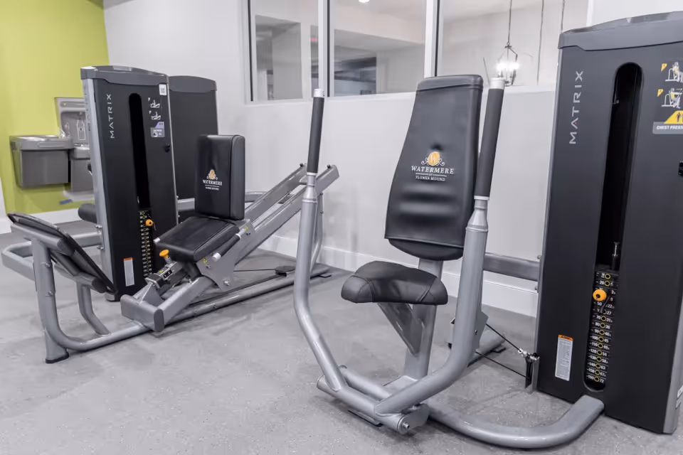 Indoor fitness room with exercise machines branded with Watermere at Flower Mound logo, including a chest press and leg press machine, set against a light gray wall and a green accent wall with a water fountain in the background.