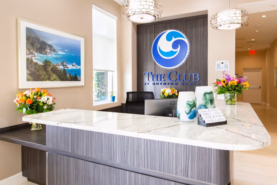 Reception desk area at The Club at Boynton Beach with a marble countertop, two modern pendant lights, flower arrangements, a computer monitor, and a large logo sign on the wall behind the desk. A framed coastal landscape photograph is on the wall to the left, and a hallway extends to the right.