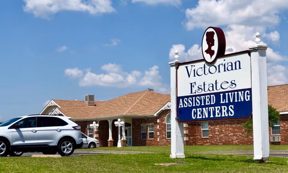 Exterior view of Victorian Estates Assisted Living Centers building with a large sign in the foreground and a white SUV parked on the driveway. The building has a brick facade and a brown shingled roof under a partly cloudy blue sky.