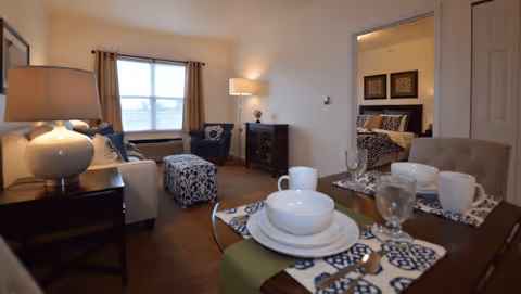 Cozy furnished living area with a set dining table in the foreground, sofa, armchair and ottoman, and a bedroom visible through an open doorway.