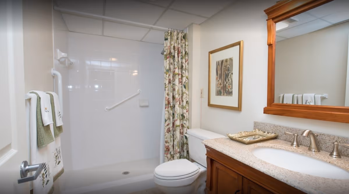 A clean and well-lit bathroom featuring a walk-in shower with a floral shower curtain, a white toilet, a wooden vanity with a granite countertop and an oval sink, a large wooden framed mirror above the sink, and a set of green and white towels hanging on a rack next to the shower.
