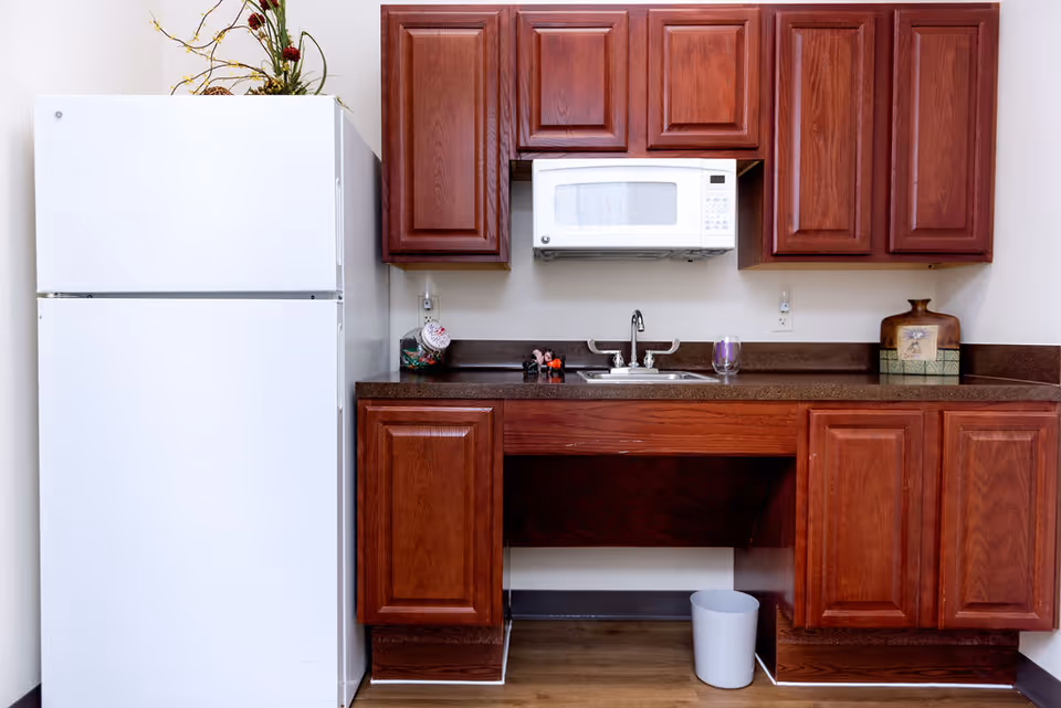 A kitchen area with a white refrigerator on the left, dark wood cabinets above and below a brown countertop. A white microwave is mounted above a stainless steel sink with a faucet. There are decorative items on the countertop including a glass jar, small figurines, a purple glass, and a ceramic container. A small white trash bin is on the floor under the counter.