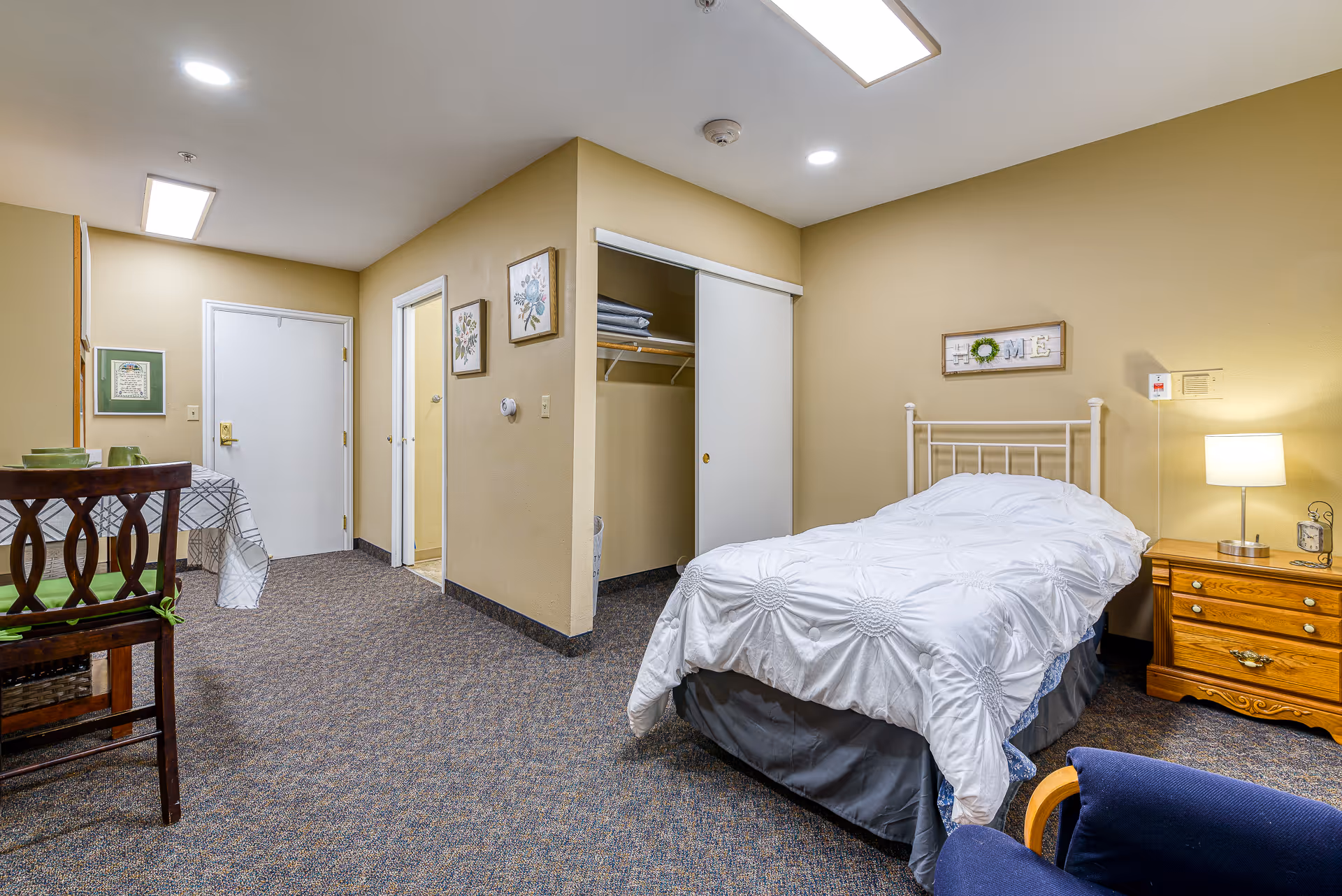 A cozy assisted living bedroom with a single bed covered in a white comforter, a wooden nightstand with a lamp and clock, an open closet with shelves and hanging space, a small dining table with chairs, and beige walls decorated with framed floral artwork and a 'HOME' sign above the bed.