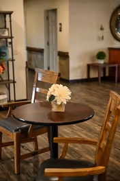 A round wooden table with a small flower arrangement in a vase at the center, surrounded by wooden chairs with dark cushions, set on a wooden floor in a warmly lit interior space with a hallway and furniture in the background.