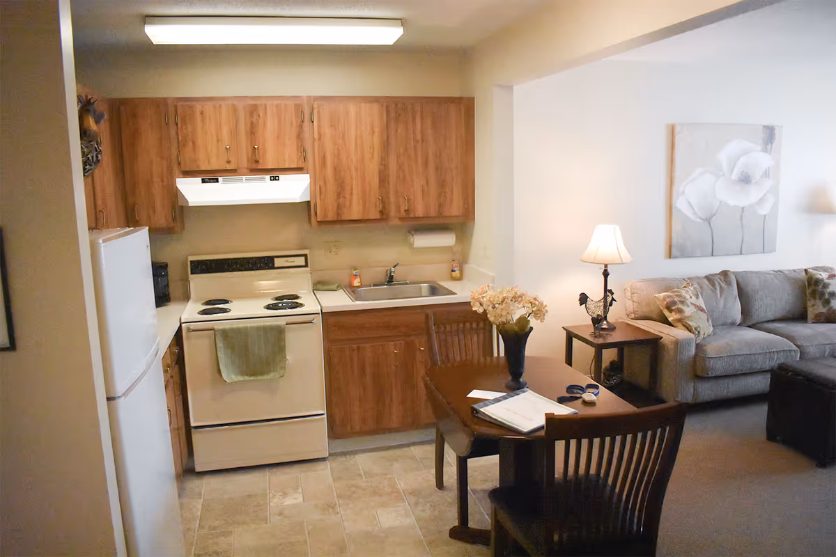 A small kitchen area with wooden cabinets, a white stove with a green towel hanging on the handle, a white refrigerator, and a sink. Adjacent to the kitchen is a dining table with two wooden chairs, a vase with flowers, and some papers on the table. In the background, there is a living room area with a beige couch, a side table with a lamp, and a wall painting of white flowers.