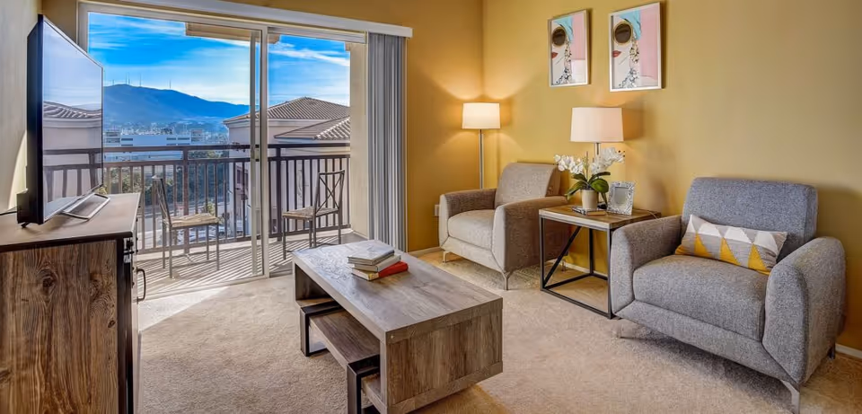 Sunlit living room with two gray armchairs, a wooden coffee table and TV, opening to a balcony with mountain views.