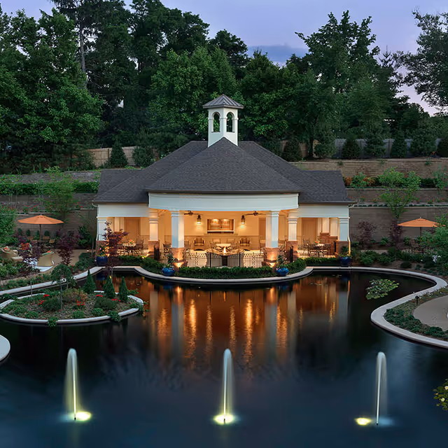 A pavilion-style building lit at dusk sits behind a reflecting pond with three illuminated fountains and landscaped gardens.