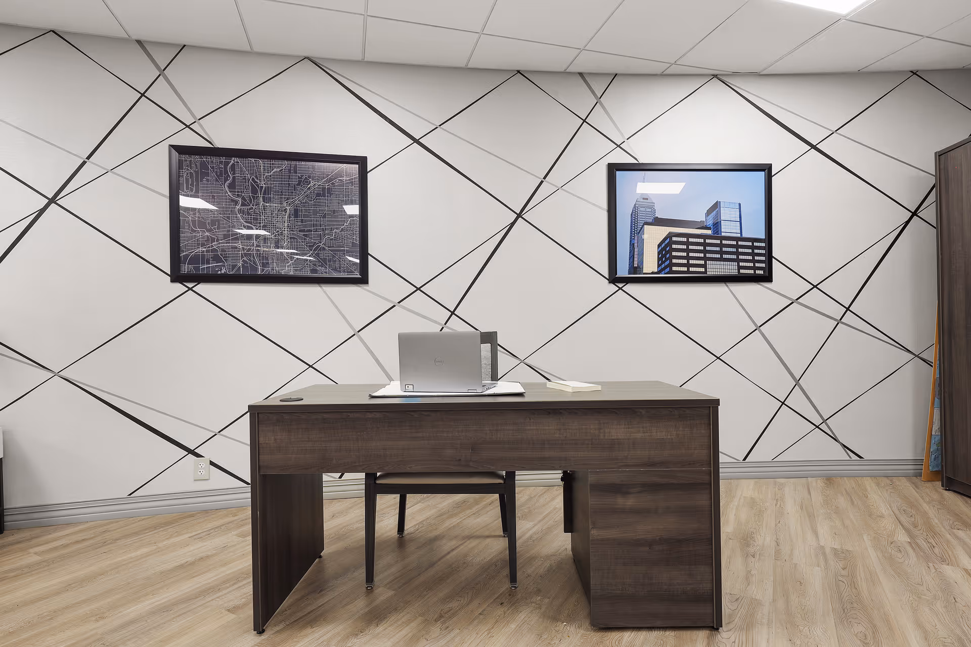 Modern office room with a wooden desk and laptop against a geometric-patterned wall with two framed pictures.