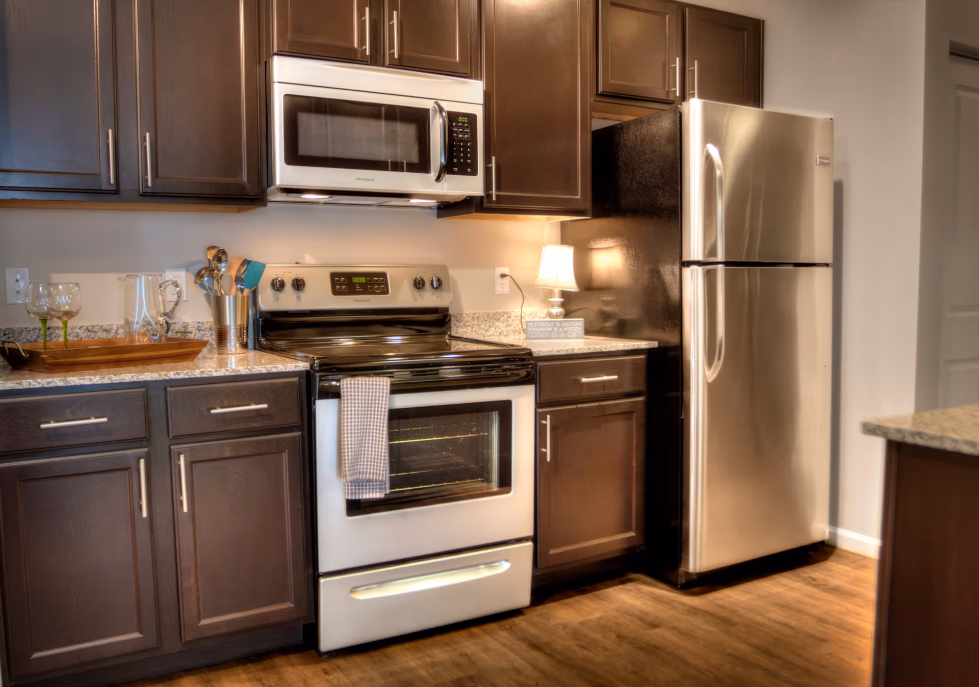 Modern kitchen with dark wooden cabinets, a stainless steel refrigerator, a stove with an oven, a microwave above the stove, and a countertop with a small lamp, utensils, and glassware.