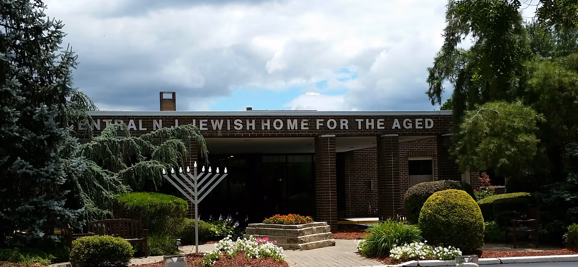 Exterior view of a brick building with the sign 'Central N. Jewish Home for the Aged' above the entrance. The foreground features a landscaped garden with trimmed bushes, flowers, benches, and a large menorah. Trees frame the scene under a partly cloudy sky.