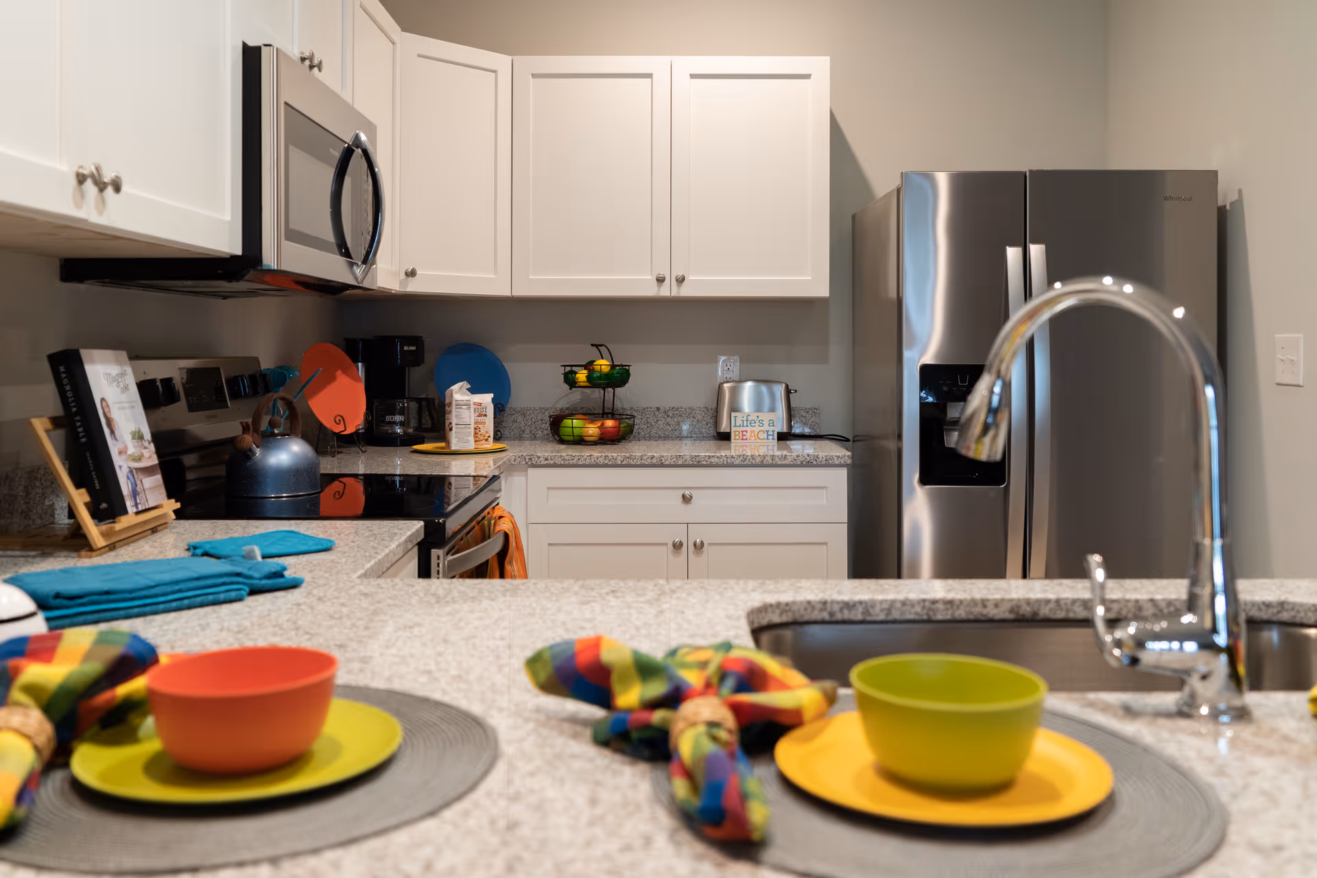 Modern kitchen with white cabinets, stainless steel refrigerator and microwave, granite countertops, a sink with a curved faucet, colorful bowls and plates on the counter, a kettle on the stove, a coffee maker, and a two-tiered fruit basket.