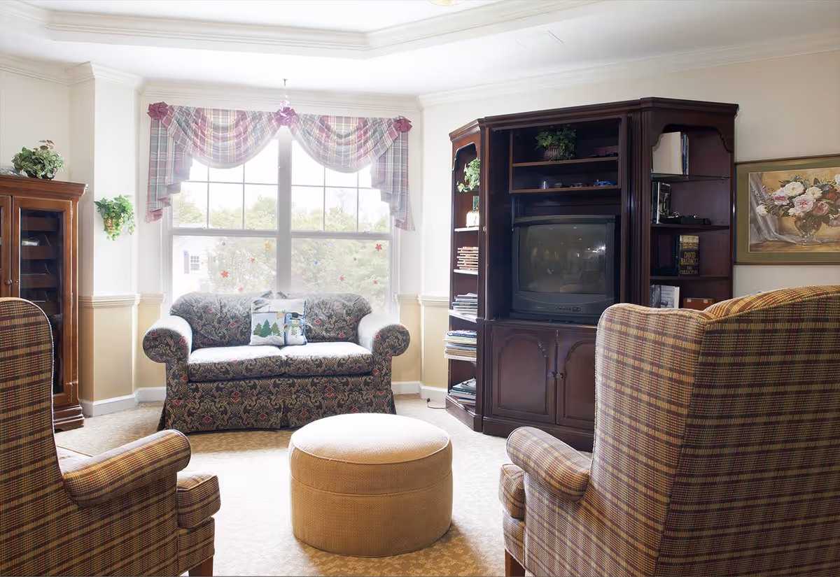 Bright living room with a patterned loveseat and two plaid armchairs arranged around a round ottoman facing a wooden entertainment center beneath a large window with plaid valances.