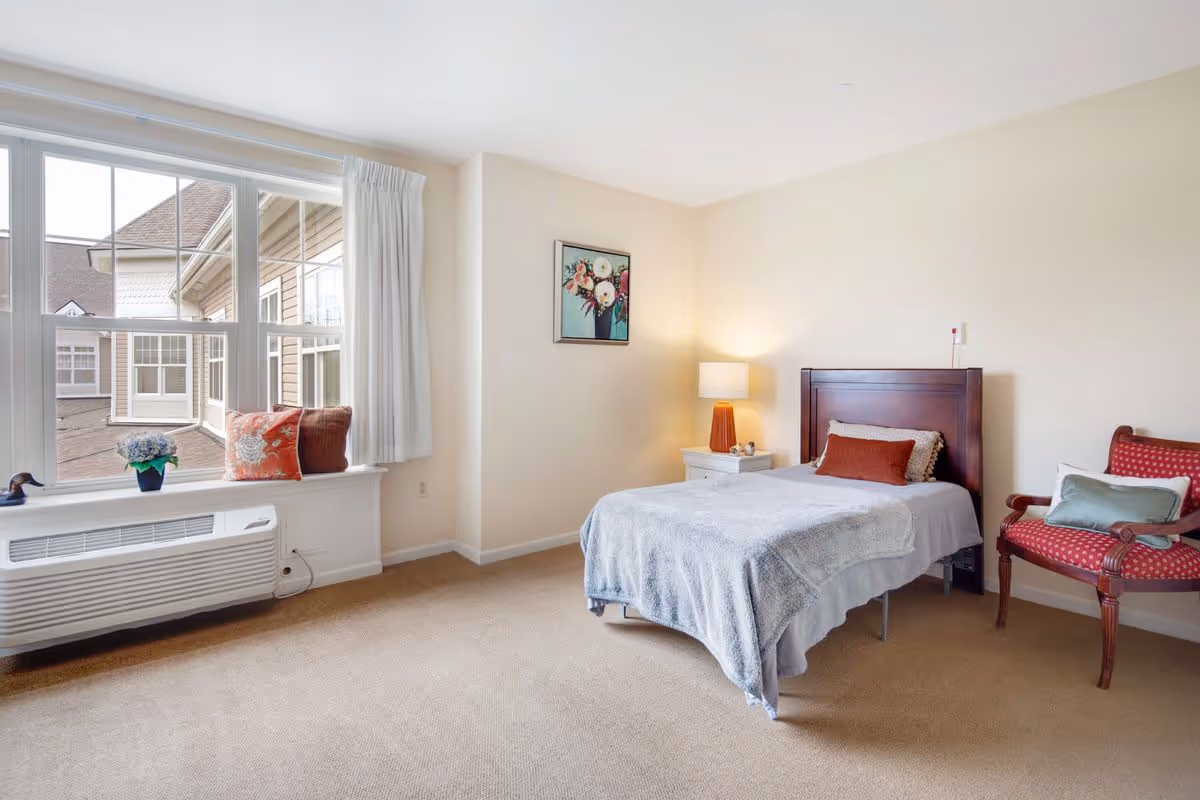 A bright bedroom with a single bed covered in a gray blanket and orange pillows. Next to the bed is a white nightstand with a lamp and decorative items. A red upholstered chair with a green pillow is placed near the bed. Large windows with white curtains let in natural light, and a small plant and decorative pillow sit on the windowsill. A framed floral painting hangs on the beige wall.