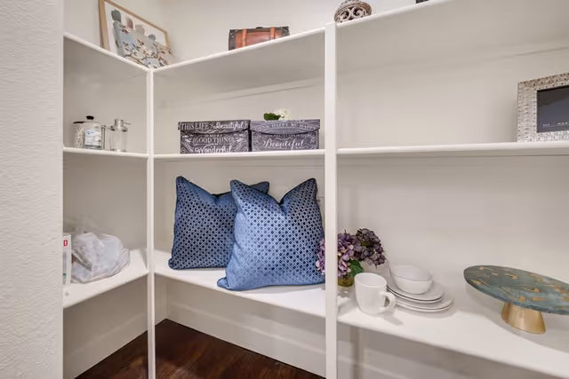 White built-in shelving unit with decorative items including two blue patterned pillows, stacked plates and bowls, a white mug, a small bouquet of purple flowers, a decorative cake stand, framed pictures, and small storage boxes with inspirational text.