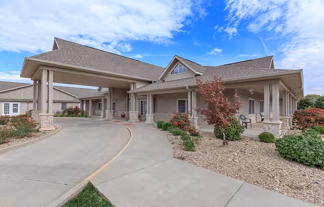 Exterior view of a senior living facility building with a covered entrance driveway, beige siding, and a landscaped area with bushes and small trees under a partly cloudy sky.