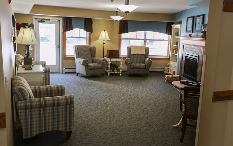 A cozy senior living facility common area with four striped armchairs arranged around a small white table. The room features large windows with brown valances, a fireplace with a mantel decorated with framed pictures, and a side table with a lamp. The carpet is patterned and the walls are painted in warm tones.