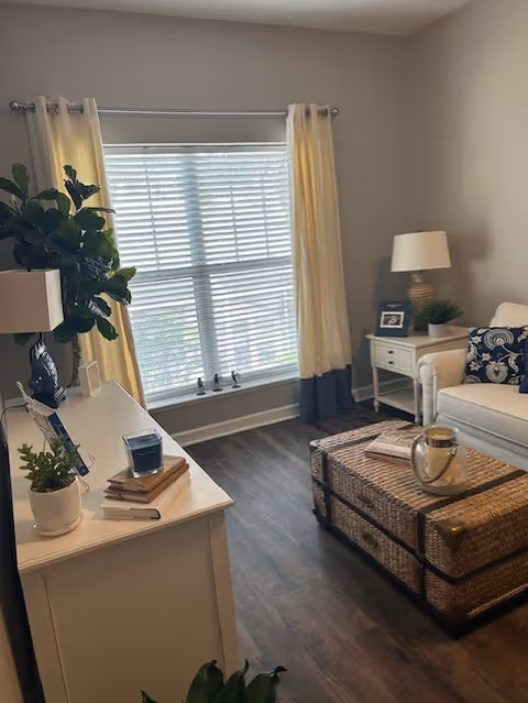 Cozy living room with a large window, white sofa, wicker trunk coffee table, side table with lamp, and decorative plants.