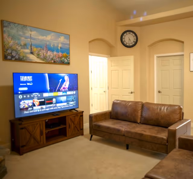 A cozy living room with a brown leather sofa and a flat-screen TV on a wooden stand. The TV screen displays a streaming service interface. Above the TV is a colorful painting of a seaside scene with flowers and sailboats. There are two white doors and a wall clock showing the time as 10:10 on the beige walls.