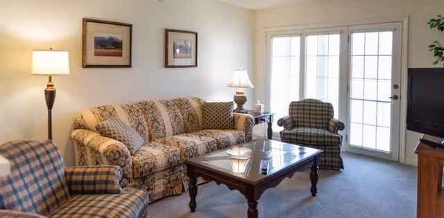 Sunlit living room with a patterned sofa and two plaid armchairs around a wooden coffee table in front of glass French doors.