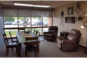 A cozy interior common area with a wooden dining table surrounded by six chairs, two brown recliner chairs, and large windows with blinds partially drawn, overlooking a parking lot outside. The walls are decorated with framed pictures and a decorative windmill piece.