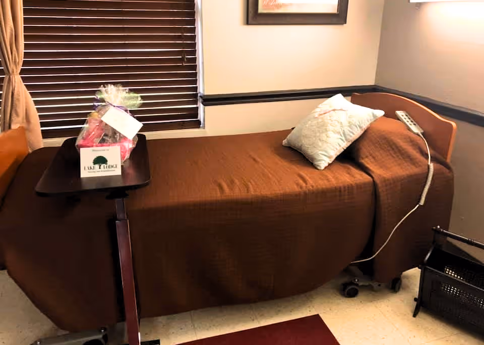 A single hospital-style bed with a brown blanket and a white pillow in a room with beige walls and a window with closed brown blinds. A small overbed table holds a gift basket with a card that reads 'Lake Lodge Nursing & Rehabilitation.' A remote control is attached to the bed, and a black basket is on the floor nearby.