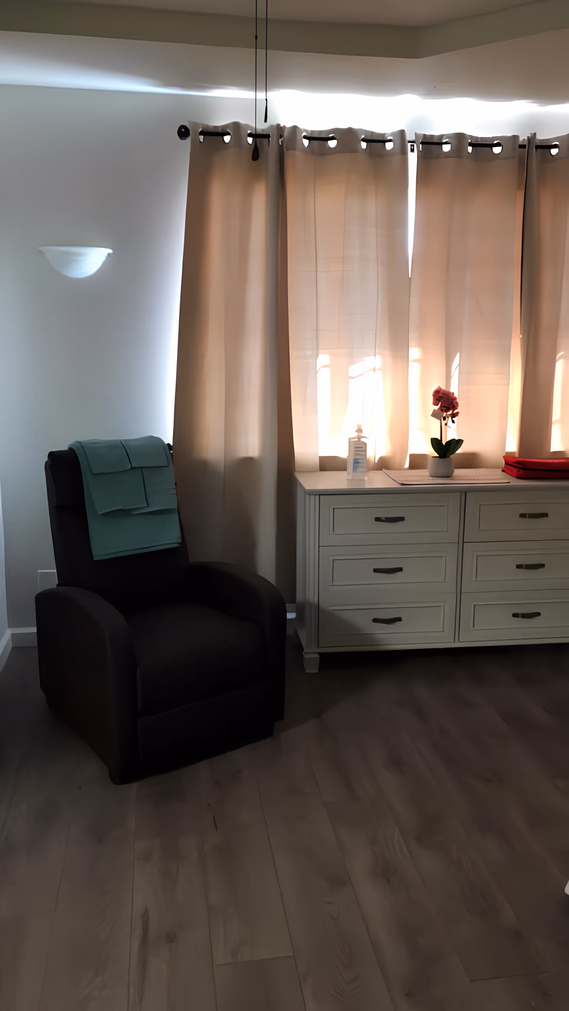 A cozy corner of a room featuring a dark recliner chair with folded green towels draped over the backrest, next to a white dresser with six drawers. On top of the dresser, there is a small potted pink orchid and a bottle of hand sanitizer. Beige curtains cover the window behind the dresser, and a wall-mounted light fixture is visible on the left wall. The floor is made of wood.