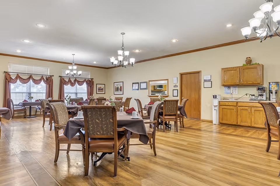 A spacious dining room with multiple tables covered in brown tablecloths, each set with white cups and saucers. The room features wooden chairs with patterned upholstery, large windows with red valance curtains, wooden flooring, and ceiling chandeliers. On the right side, there is a wooden cabinet with a coffee machine and water dispenser.