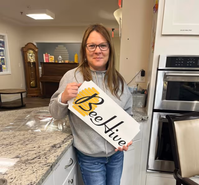 A woman with glasses and long hair stands in a kitchen area holding a white sign with the text 'BeeHive' and a yellow bee hive graphic. The kitchen has granite countertops, white cabinets, and a built-in oven. In the background, there is a wooden grandfather clock, a piano, and some decorative items on a shelf.