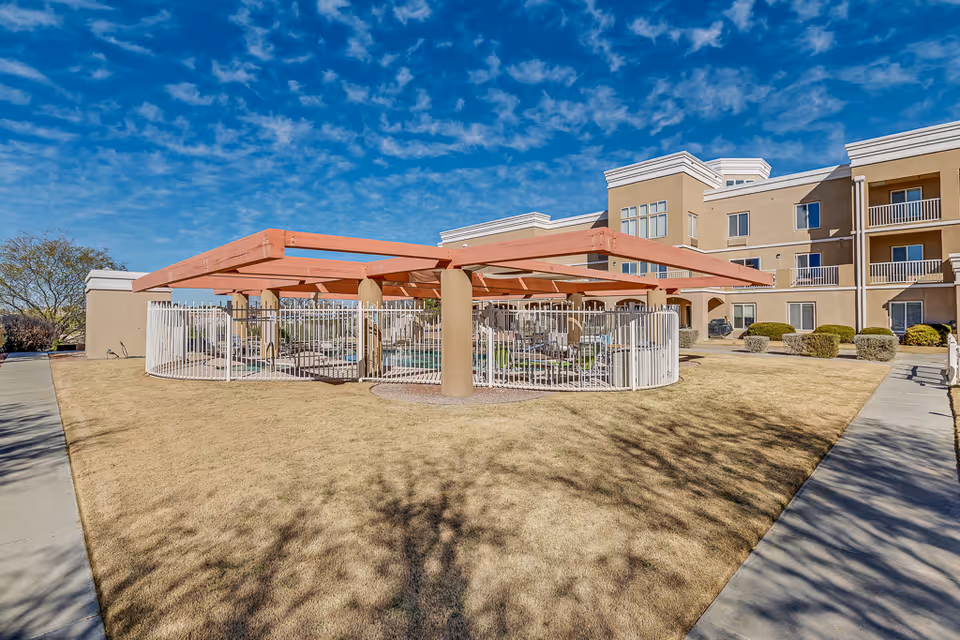 Outdoor courtyard with a fenced circular pergola and seating in front of a three-story senior living building under a blue sky.