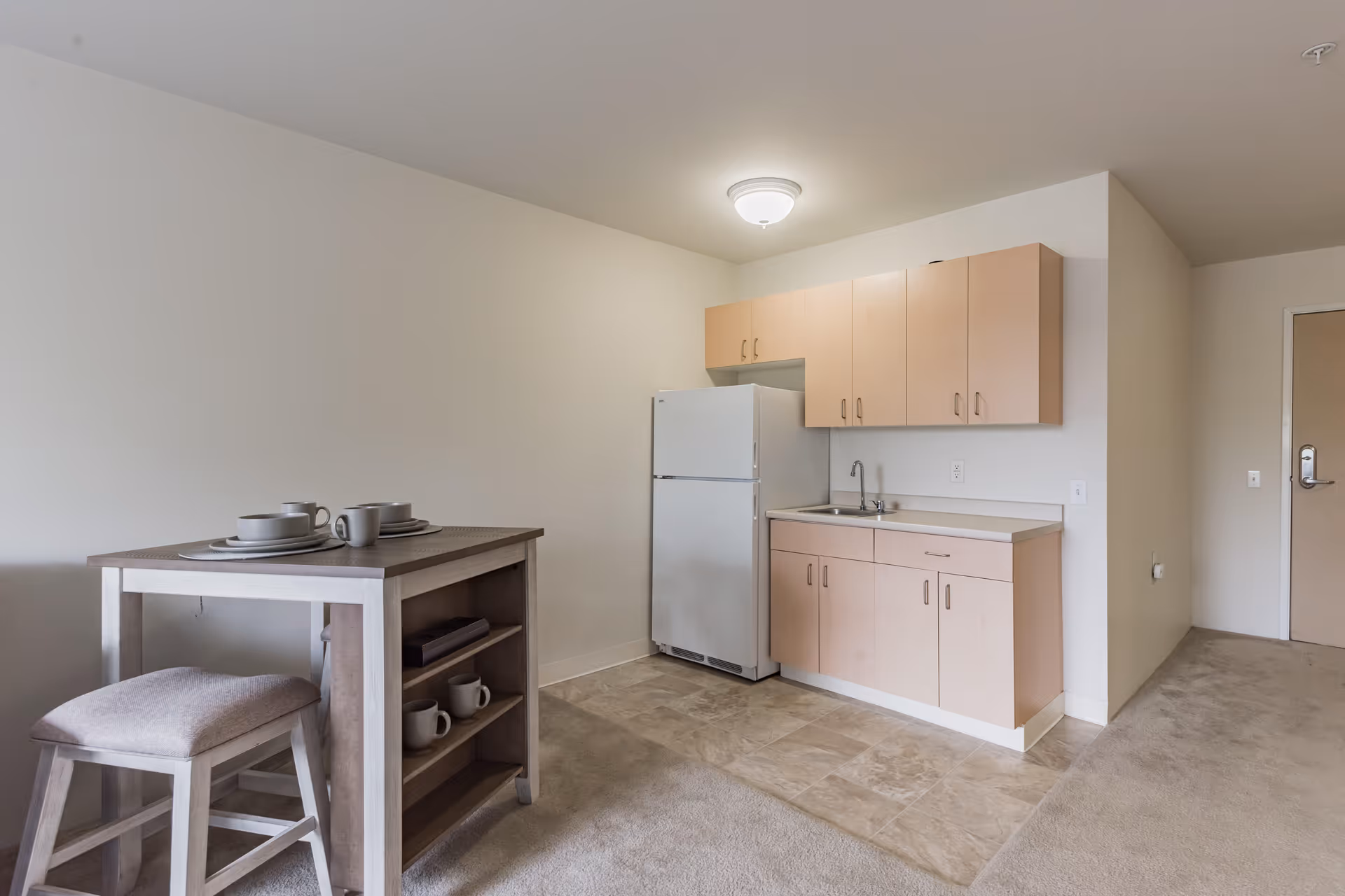 Small kitchen area in a senior living facility with light wood cabinets, a white refrigerator, a sink, and a small dining table with two place settings and a cushioned stool. The floor transitions from tile in the kitchen area to carpet in the adjacent space.