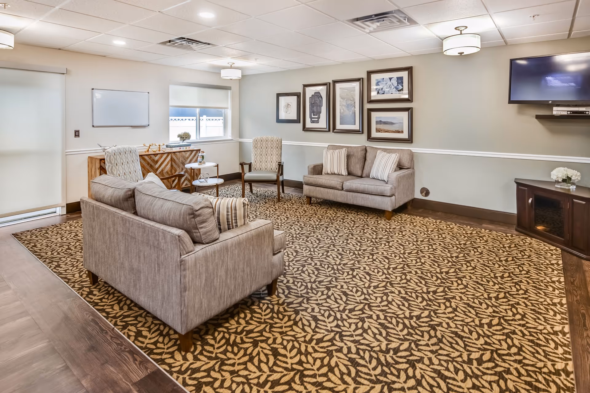 A cozy living room area with two beige sofas, two patterned armchairs, a patterned carpet, and framed artwork on the wall. There is a flat-screen TV mounted on the wall above a dark wooden cabinet with a small flower arrangement. The room has a window with a roller shade and ceiling lights.