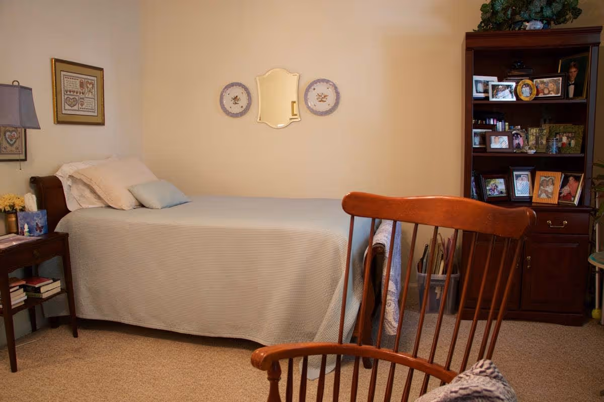 A cozy bedroom with a single bed covered in a light blue bedspread and several pillows. There is a wooden rocking chair in the foreground, a small side table with books and a lamp on the left, and a wooden bookshelf filled with framed photos and decorative items on the right. The walls are decorated with a mirror and two decorative plates.