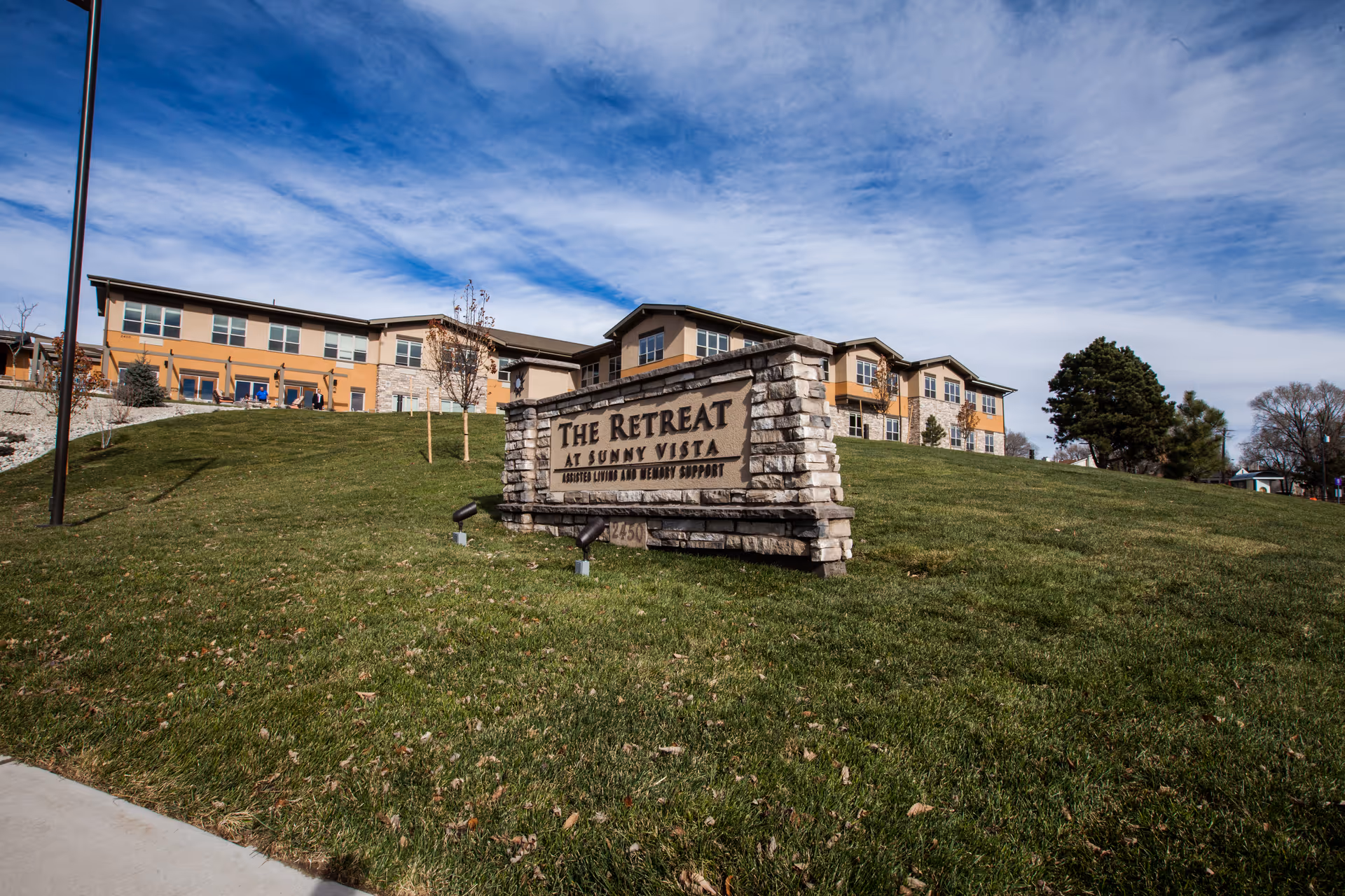 Stone sign reading "The Retreat at Sunny Vista" on a grassy hill in front of a two-story senior living building under a partly cloudy sky.