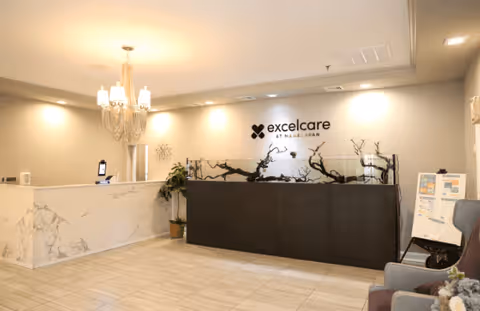 Reception area of Accela Rehab and Care Center at Manalapan featuring a marble reception desk, a chandelier light fixture, a decorative black planter with branches, a potted plant, and seating area with chairs. The wall behind the planter displays the logo and name 'excelcare at manalapan'.