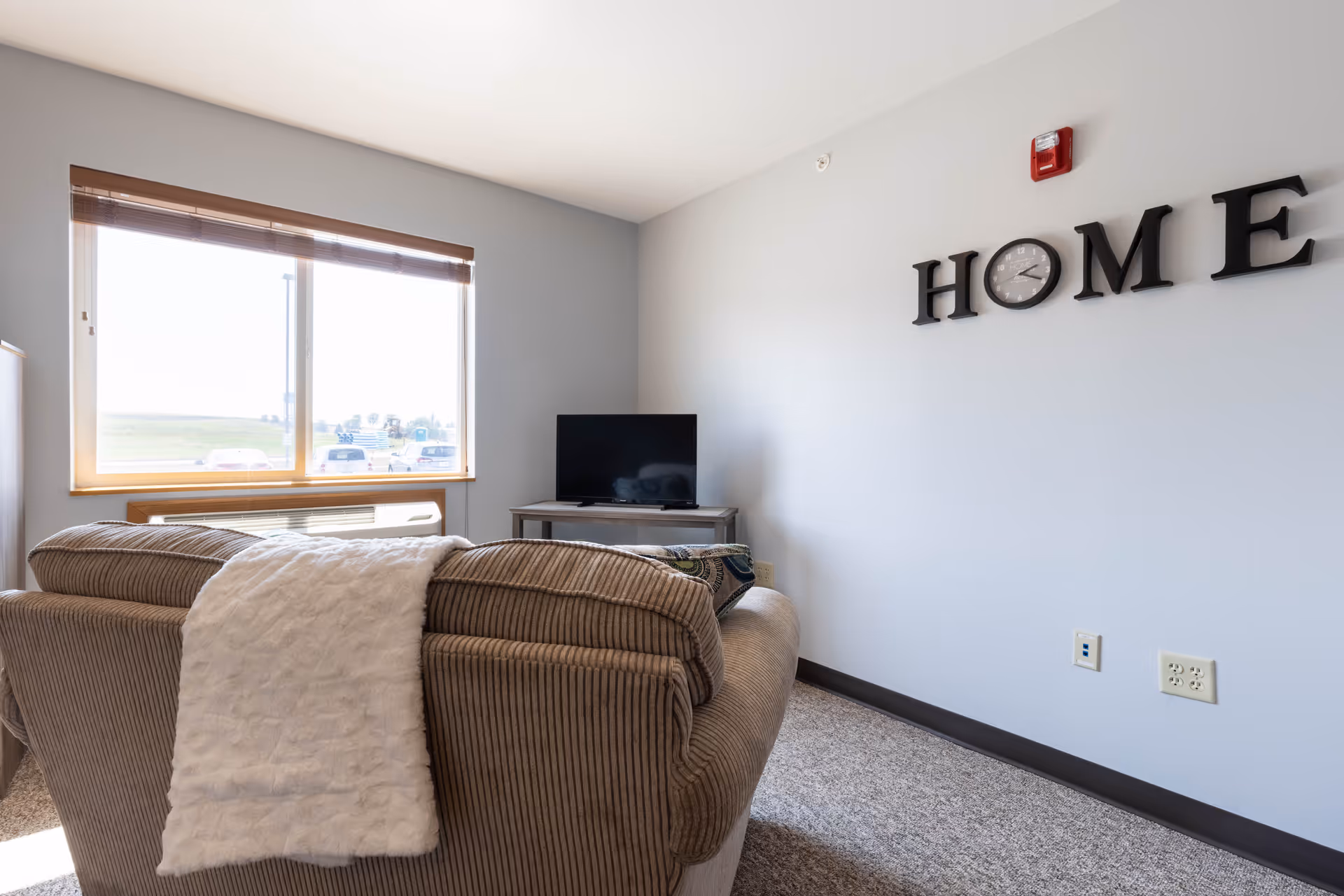 A cozy living room with a large window letting in natural light, a brown corduroy sofa with a white throw blanket draped over the back, a small TV on a stand against a light gray wall, and the word 'HOME' displayed on the wall with the letter 'O' replaced by a clock.