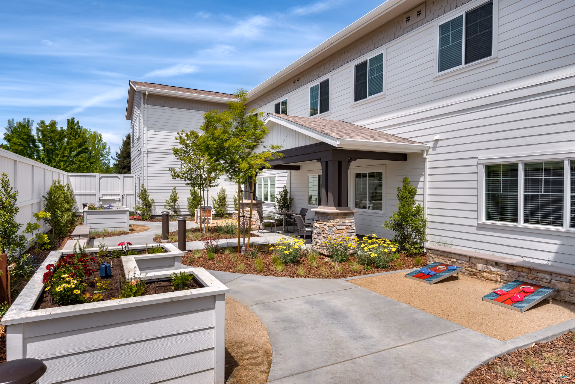 Outdoor garden area at Clearwater at Sonoma Hills featuring raised flower beds with blooming flowers, a paved walkway, small trees, and a covered seating area with chairs. There is also a cornhole game set up on a sandy patch near the building with white siding and multiple windows.