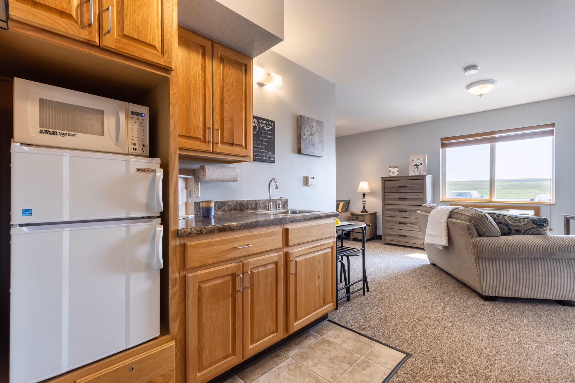 Interior view of a senior living facility unit showing a small kitchen area with wooden cabinets, a white microwave, and a refrigerator on the left. The kitchen counter has a sink and a paper towel holder. Beyond the kitchen is a living area with a beige couch, a wooden dresser, a side table with a lamp, and a large window letting in natural light.