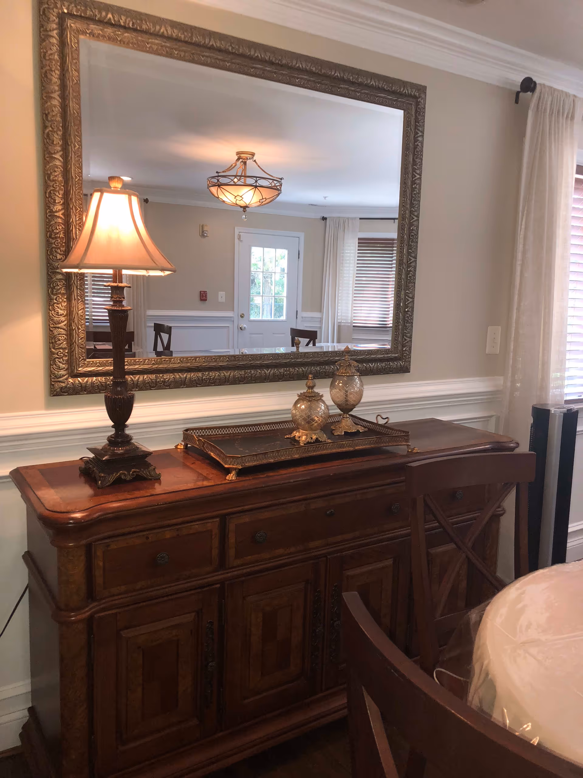 A wooden sideboard with ornate detailing topped with a decorative tray holding two glass jars and a classic table lamp with a beige shade. Above the sideboard is a large, ornate framed mirror reflecting a door, windows with blinds, and part of a dining table with chairs. The room has beige walls with white wainscoting and crown molding, and sheer curtains on the windows.