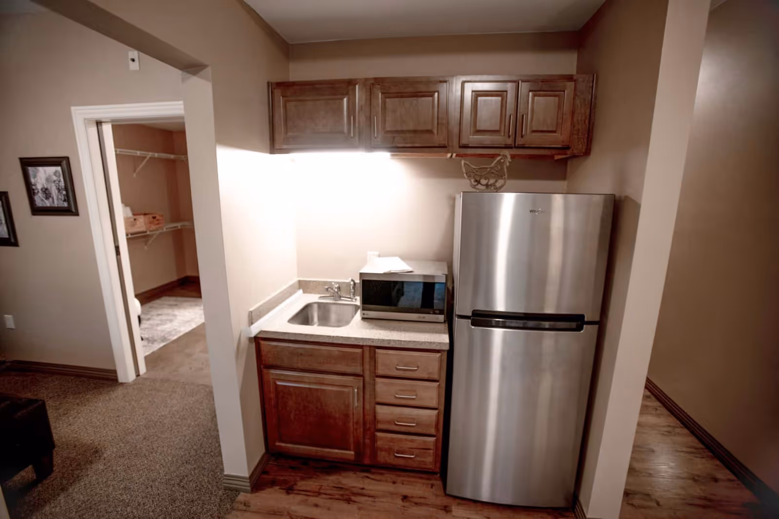 Small kitchenette area with wooden cabinets, a stainless steel refrigerator, a microwave on the countertop, and a small sink. Adjacent to the kitchenette is an open doorway leading to a walk-in closet with shelving. The floor is a combination of wood and carpet.